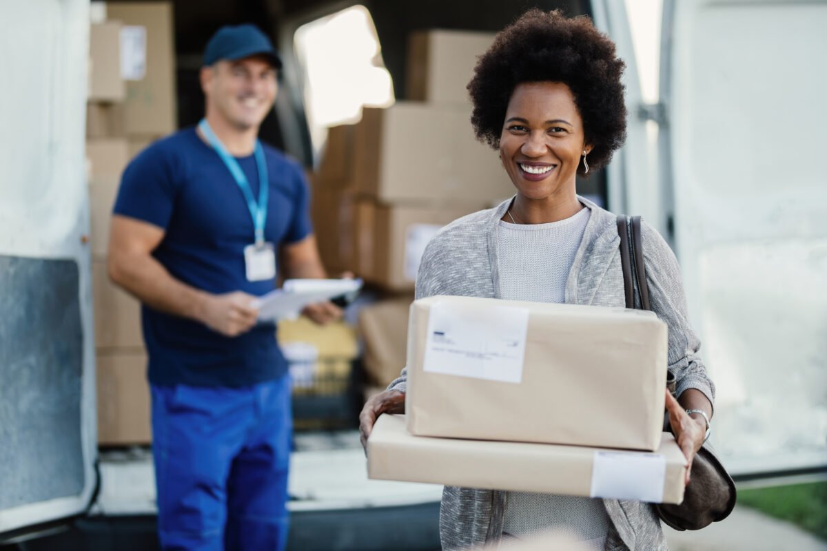 Portrait of happy black woman feeling satisfied with package delivery ad holding boxes while looking at camera. The courier is in the background.